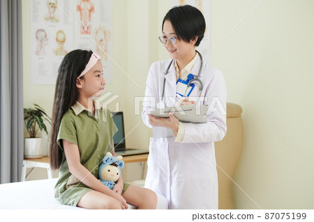 Asian doctor in white coat making notes in medical card while talking to little patient at hospital 87075199