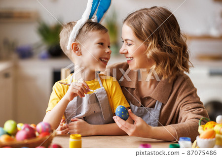 Loving young mother teaching happy little kid son to decorate Easter eggs while sitting in kitchen 87075486