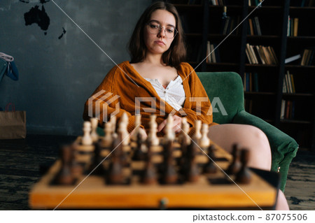 Portrait of thoughtful young woman in elegant eyeglasses thinking about chess move sitting in armchair in dark library room. Pretty intelligent lady playing logical board game alone at home. 87075506