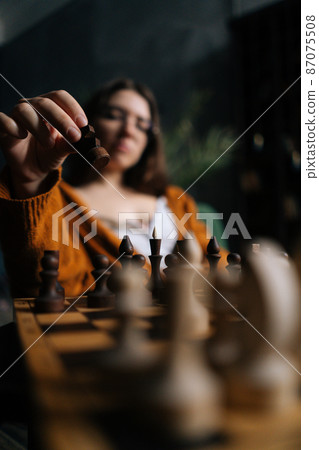 Vertical selective focus shot of confident businesswoman in elegant eyeglasses making chess move sitting on armchair in dark room. Pretty intelligent lady playing logical board game alone at home. Vertical selective focus shot of confident businesswoman in elegant eyeglasses making chess move sitting on armchair in dark room. Pretty intelligent lady playing logical board game alone at home. 87075508