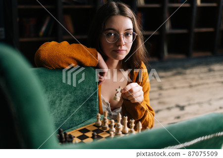 Portrait of attractive young woman wearing elegant eyeglasses making chess move with knight piece sitting in floor, looking at camera. Pretty intelligent lady playing logical board game alone at home. Portrait of attractive young woman wearing elegant eyeglasses making chess move with knight piece sitting in floor, looking at camera. Pretty intelligent lady playing logical board game alone at home. 87075509