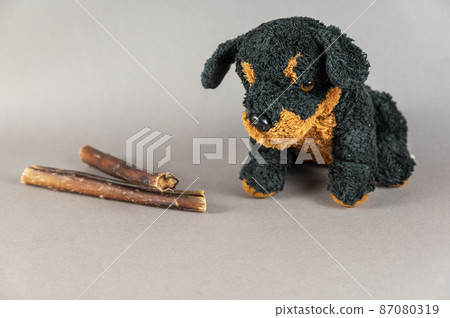 Pet treats and a children's toy opposite the gray background. Natural dried treats and a black stuffed dog. Bully sticks. Pet supplies. 87080319