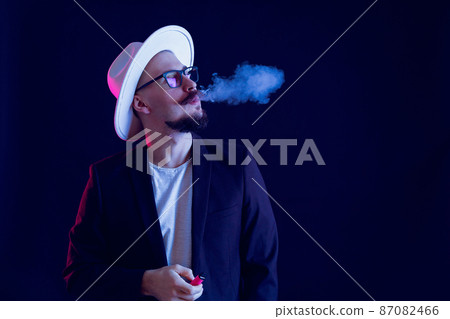 Hipster beard man in white wide-brimmed hat holds vape and smoking on dark background. Color filters  87082466