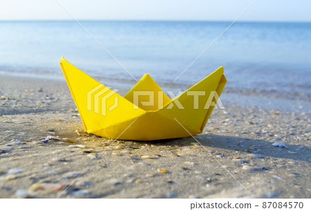 Small paper yellow boat on sand near water on background of sea waves Small paper yellow boat on sand near water on background of sea waves 87084570