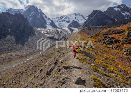 Traveling man in red with big orange backpack go on way to high mountain ridge along a narrow path. Dramatic landscape with a tourist high in the mountains. Traveling man in red with big orange backpack go on way to high mountain ridge along a narrow path. Dramatic landscape with a tourist high in the mountains. 87085473