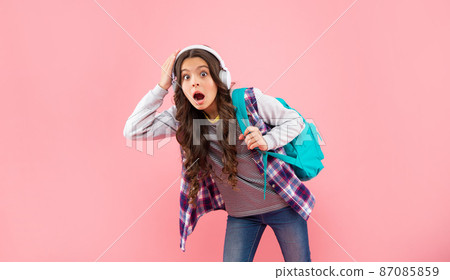 shocked teen girl with backpack wearing headphones on pink background, childhood 87085859