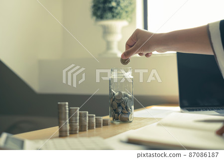 Woman's hand. Put a coin into a glass bottle with coins, saving money with coins, stepping into a successful growing business, and saving for thought in retirement. Retirement savings concept Woman's hand. Put a coin into a glass bottle with coins, saving money with coins, stepping into a successful growing business, and saving for thought in retirement. Retirement savings concept 87086187
