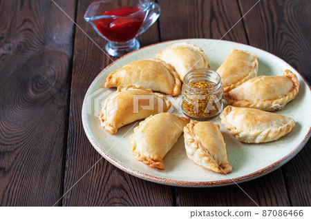 Freshly baked small empanadas on a plate with ketchup and mustard on a wooden background. Freshly baked small empanadas on a plate with ketchup and mustard on a wooden background. 87086466