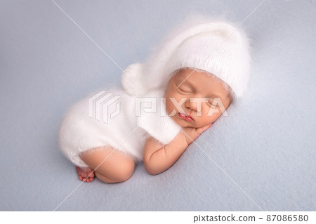 Sleeping newborn girl in the first days of life in a white soft bodysuit with a knitted woolen white cap on a blue background. Studio macro photography, portrait of a newborn. Woman's happiness. Sleeping newborn girl in the first days of life in a white soft bodysuit with a knitted woolen white cap on a blue background. Studio macro photography, portrait of a newborn. Woman's happiness. 87086580