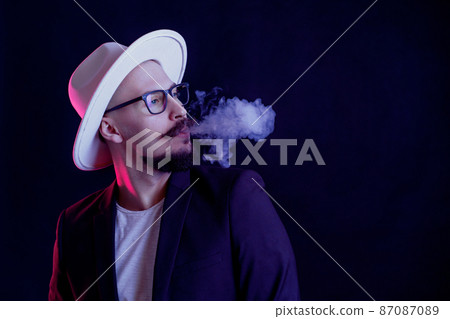 Portrait of beard man in white wide-brimmed hat who blows smoke on dark background. Color filters 87087089