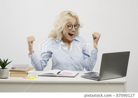 Overjoyed female entrepreneur using laptop and gesturing yes, celebrating business success, sitting at workplace 87090127