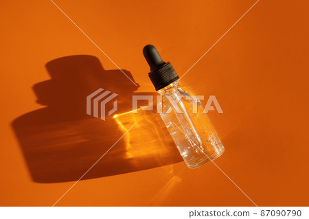 Transparent glass dropper bottle with air bubbles on orange background in sunlight. Pipette with fluid hyaluronic acid, serum, retinol. Cosmetics and healthcare concept Flat lay. Luxury beauty product 87090790