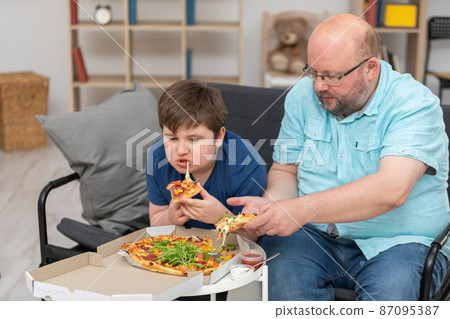 Dad and son are sitting on the sofa in the room eating pizza. Dad and son are sitting on the sofa in the room eating pizza. 87095387
