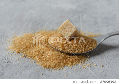 Brown cane sugar crystals and cube of refined sugar in a spoon on grey table. 87096398