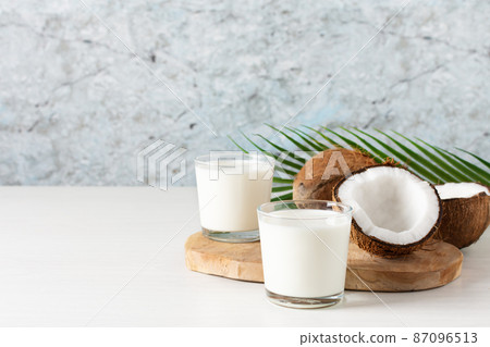 Two glasses of coconut vegan milk with coconut halves on white wooden table. 87096513