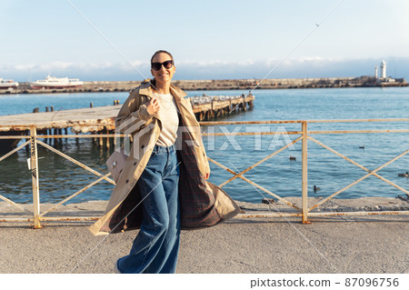 Young adult trendy stylish beautiful caucasian happy smiling woman enjoy walking by Yalta sea embankment on warm sunny day. Female person portrait wear jeans biege trench coat on urban city street 87096756