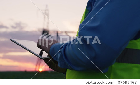 electrician with tablet hands at sunset.high power lines. electrical engineer helmet works with digital tablet. engineering business concept. electrical power distribution industry. power engineer 87099468