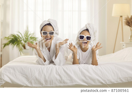 Fashion beauty portrait of happy mother and little daughter wearing towels on her head and fashionable glasses. Family cheerfully exclaims looking at camera having fun at home or in bright hotel room. Fashion beauty portrait of happy mother and little daughter wearing towels on her head and fashionable glasses. Family cheerfully exclaims looking at camera having fun at home or in bright hotel room. 87100561
