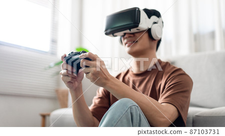 Technology Concept A man putting on a virtual reality headset and pressing a black console game while sitting on the floor Technology Concept A man putting on a virtual reality headset and pressing a black console game while sitting on the floor 87103731