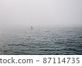 A man on a standing surf rows a paddle far out into the sea in the fog. 87114735
