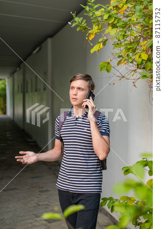 Portrait of young caucasian man in casual clothes standing outdoors and making phone call. Using smartphone, talking by the phone. Urban lifestyle. 87115752