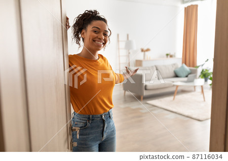 Cheerful African Woman Opening Door Welcoming You Standing At Home Cheerful African Woman Opening Door Welcoming You Standing At Home 87116034