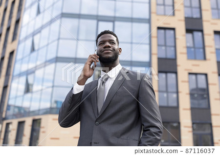 Business communications concept. Young african american businessman in suit walking outdoors, talking on cellphone 87116037
