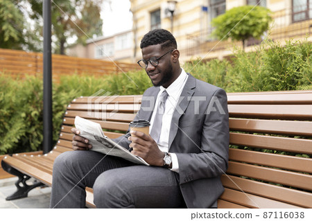 Confident african american businessman reading daily news in newspaper and drinking coffee to go, sitting on bench Confident african american businessman reading daily news in newspaper and drinking coffee to go, sitting on bench 87116038