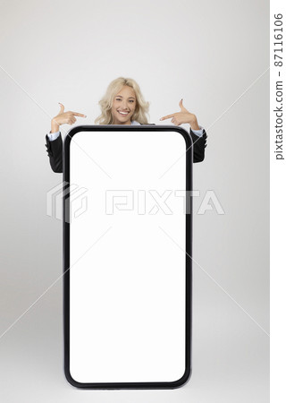 Young businesswoman standing behind huge smartphone and pointing at white empty screen, showing space for your design 87116106