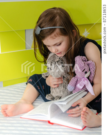 A little girl is reading a book of fairy tales to her toys in her room on the bed. A little girl is reading a book of fairy tales to her toys in her room on the bed. 87118653