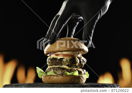 Hand putting top burger bun on a meat double cheeseburger with vegetables with sauce oozing  87120022