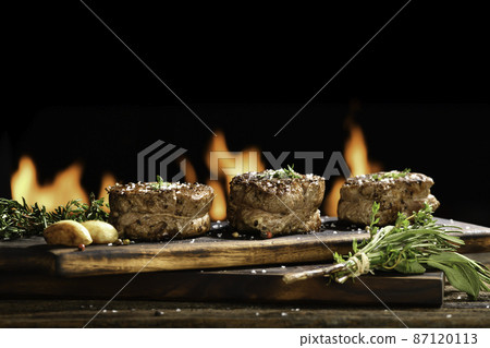 Cooked juicy steak meat beef with seasoning on wooden chopping board with flames in the background. Cooked juicy steak meat beef with seasoning on wooden chopping board with flames in the background. 87120113