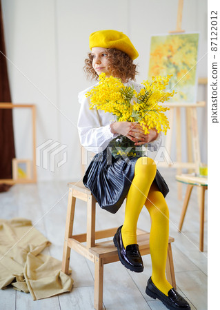 Full length of little curly girl in yellow beret who poses in studio while holding yellow flowers 87122012