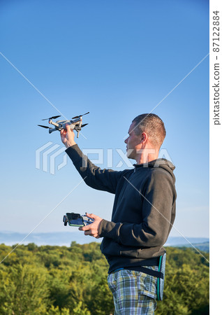Man operating drone using remote controller. Man using drone at sunset for photos and video making while standing on top of high boulder in the mountains. Man operating drone using remote controller. Man using drone at sunset for photos and video making while standing on top of high boulder in the mountains. 87122884