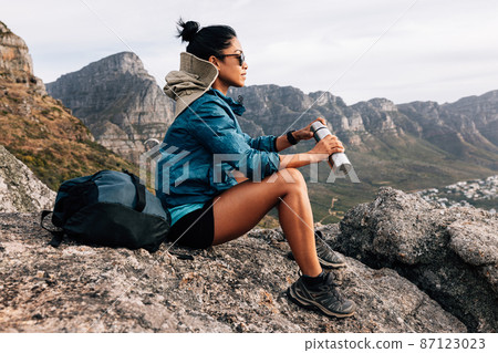 Side view of woman hiker in sports clothes enjoying the view. Female in sunglasses holding a thermos sitting on a rock and relaxing during a hike. 87123023