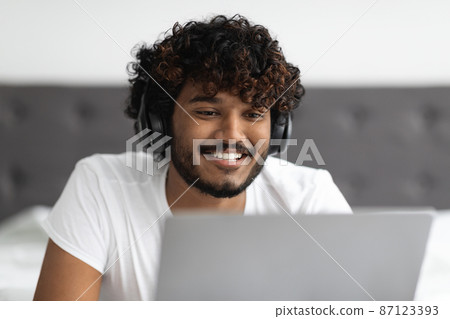 Positive millennial man using laptop and headset in bed Positive millennial man using laptop and headset in bed 87123393