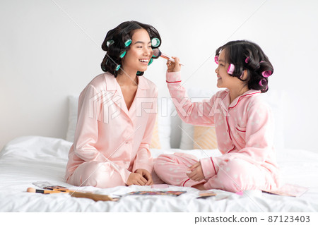 Smiling teen japanese girl in pajama and curlers applies makeup to young lady on bed in bedroom interior 87123403