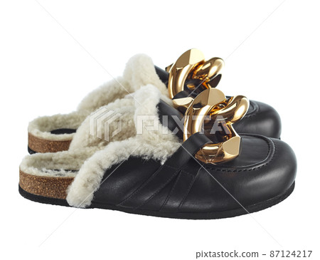 Home slippers with an open heel, made of natural sheepskin, fur inside, black leather outside, decorated with large links of a gold chain isolated on a white background. Side view. Fashionable shoes. 87124217