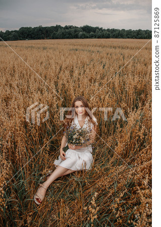 A girl sits in a field and holds a bouquet of wildflowers in her hands. Summer day. Beautiful blonde girl among the wildflowers in summer. Organic golden ripe ears of oats in field. 87125869