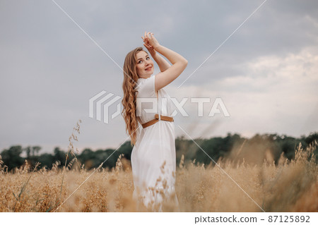 Portrait of a young woman in a white dress on a background of golden oats field. Caucasian tourist exploring rural nature with farm fields of oat cultivation, young female traveller walking. Portrait of a young woman in a white dress on a background of golden oats field. Caucasian tourist exploring rural nature with farm fields of oat cultivation, young female traveller walking. 87125892