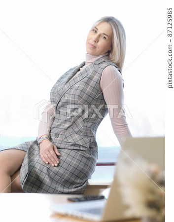 business woman having a break working indoors at office. beautiful blond woman employee sitting on a desk and getting rest. vertical shot. business woman having a break working indoors at office. beautiful blond woman employee sitting on a desk and getting rest. vertical shot. 87125985