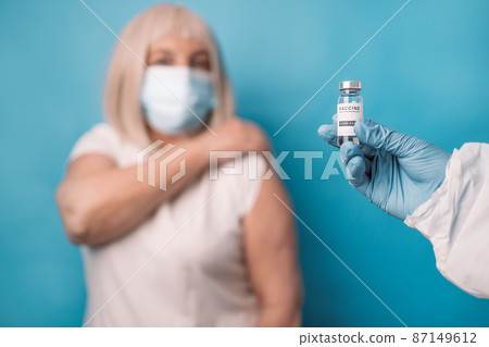 Doctor makes vaccination to senior woman, wearing face mask over blue background. Doctor makes vaccination to senior woman, wearing face mask over blue background. 87149612