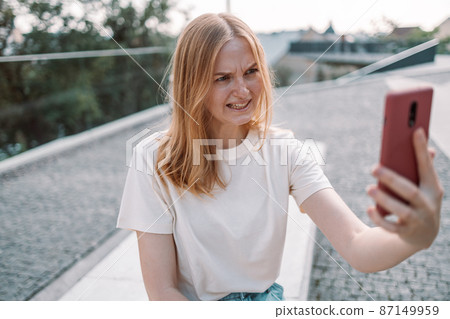 Blonde hair angry young woman posing outdoors in park take a selfie by mobile phone while standing in the park. Blonde hair angry young woman posing outdoors in park take a selfie by mobile phone while standing in the park. 87149959