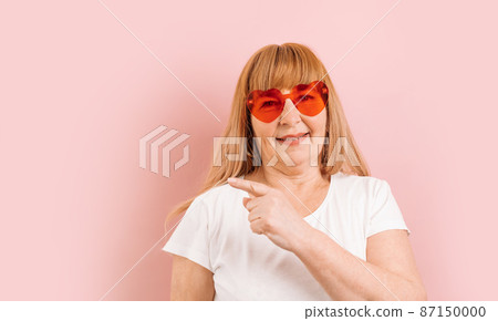 Attention promotions banner. Caucasian 50s woman in red glasses in good mood pointing finger to the side on copy space, isolated on pink background. Mock up copy space Attention promotions banner. Caucasian 50s woman in red glasses in good mood pointing finger to the side on copy space, isolated on pink background. Mock up copy space 87150000