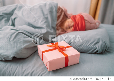Woman in red sleep mask sleeping in bed near paper gift box with satin ribbon decor. Christmas, New Year, Valentine's Day and birthday 87150012