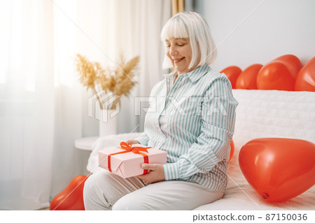 Attractive happy smiling blonde 50s woman holding pink gift box with red ribbon on 14 february sitting on a sofa at home. Attractive happy smiling blonde 50s woman holding pink gift box with red ribbon on 14 february sitting on a sofa at home. 87150036
