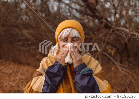 Sick adult woman coughing and blowing her nose in park during autumn season in a cold autumn day. Sick adult woman coughing and blowing her nose in park during autumn season in a cold autumn day. 87150156
