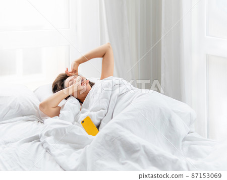 Sleepy woman is lying in bed, completely covered with white blanket. Smartphone used as alarm clock. It's hard to wake up early in morning. Woman does not get enough sleep. Sleepy woman is lying in bed, completely covered with white blanket. Smartphone used as alarm clock. It's hard to wake up early in morning. Woman does not get enough sleep. 87153069