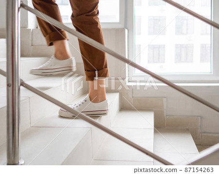 Woman in white sneakers and khaki trousers goes upstairs to her apartment. White staircase in apartment building. Casual outfit, urban fashion. 87154263