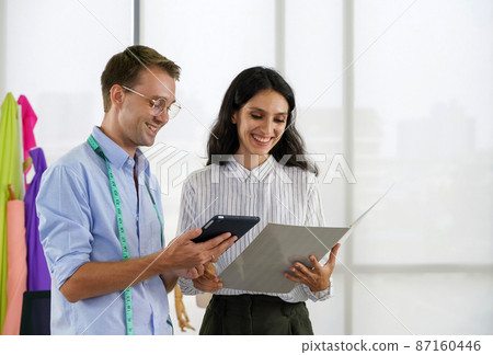 Fashion designer room, man and woman working in home base cloth design business. Young client is comparing the orders from the internet with the color charts from her catalog. 87160446